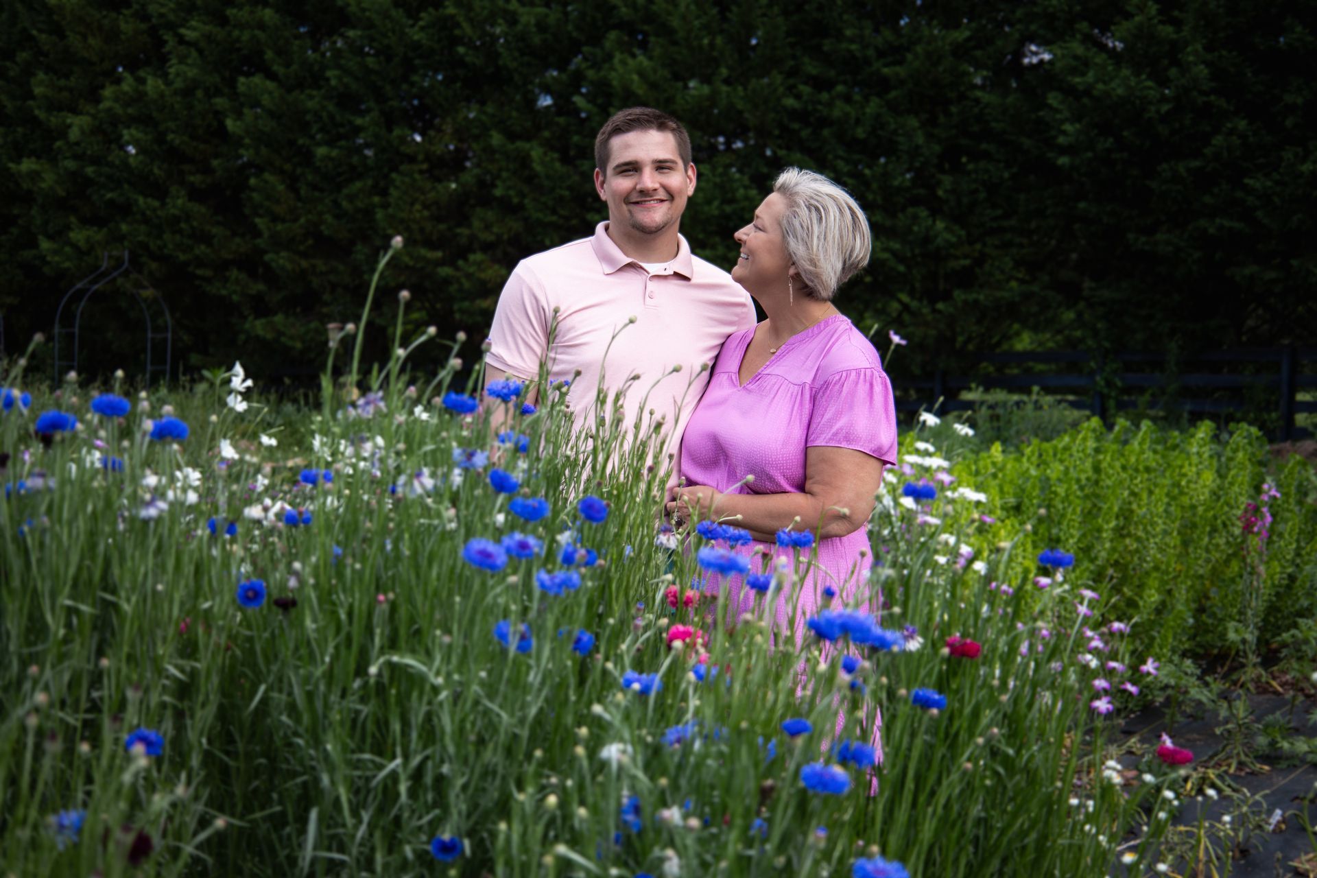 Man and woman stand in a field of blue and white flowers, smiling at each other.