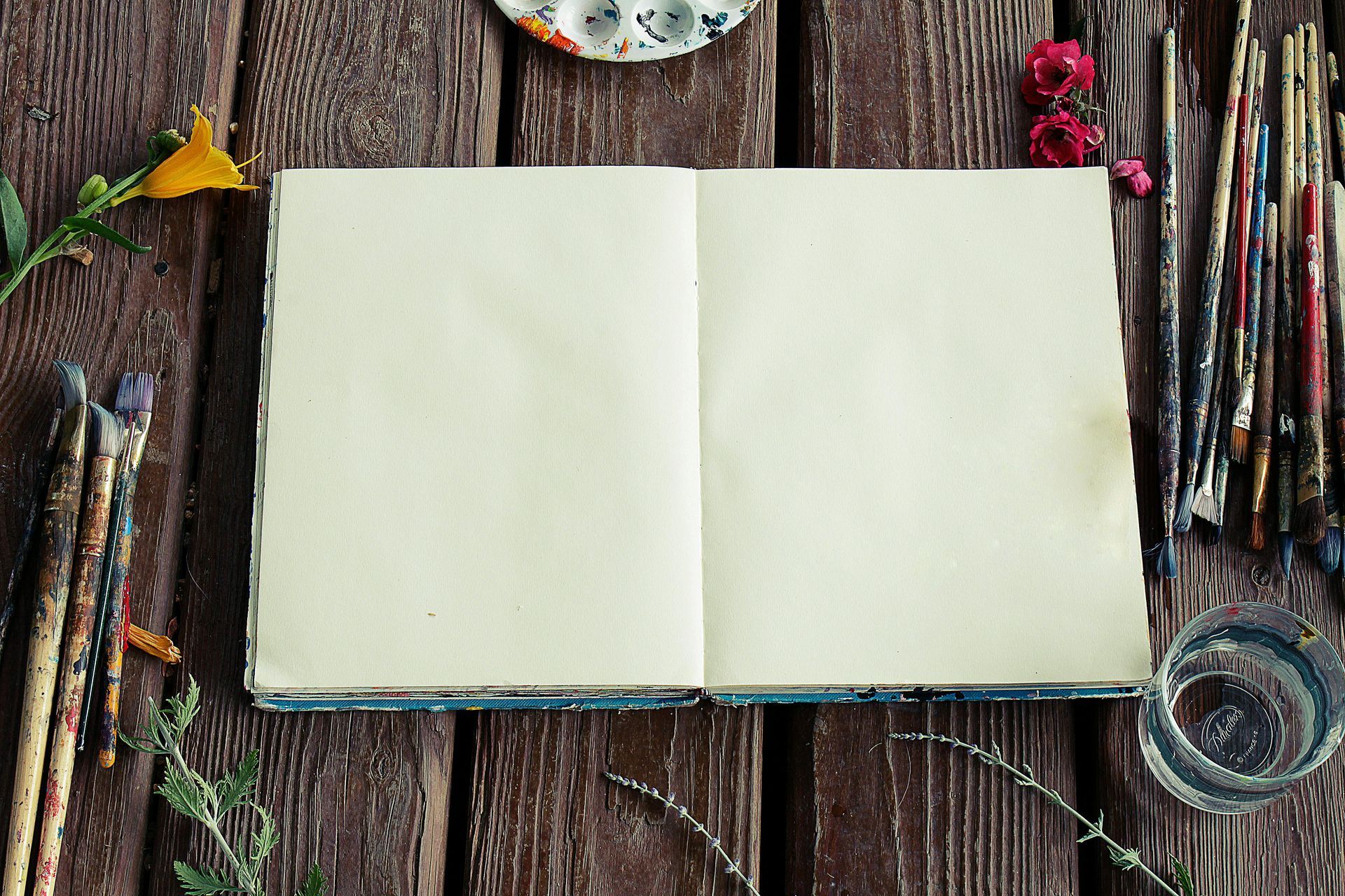 Open sketchbook on rustic wooden table with paintbrushes, palette, flowers, and water glass.