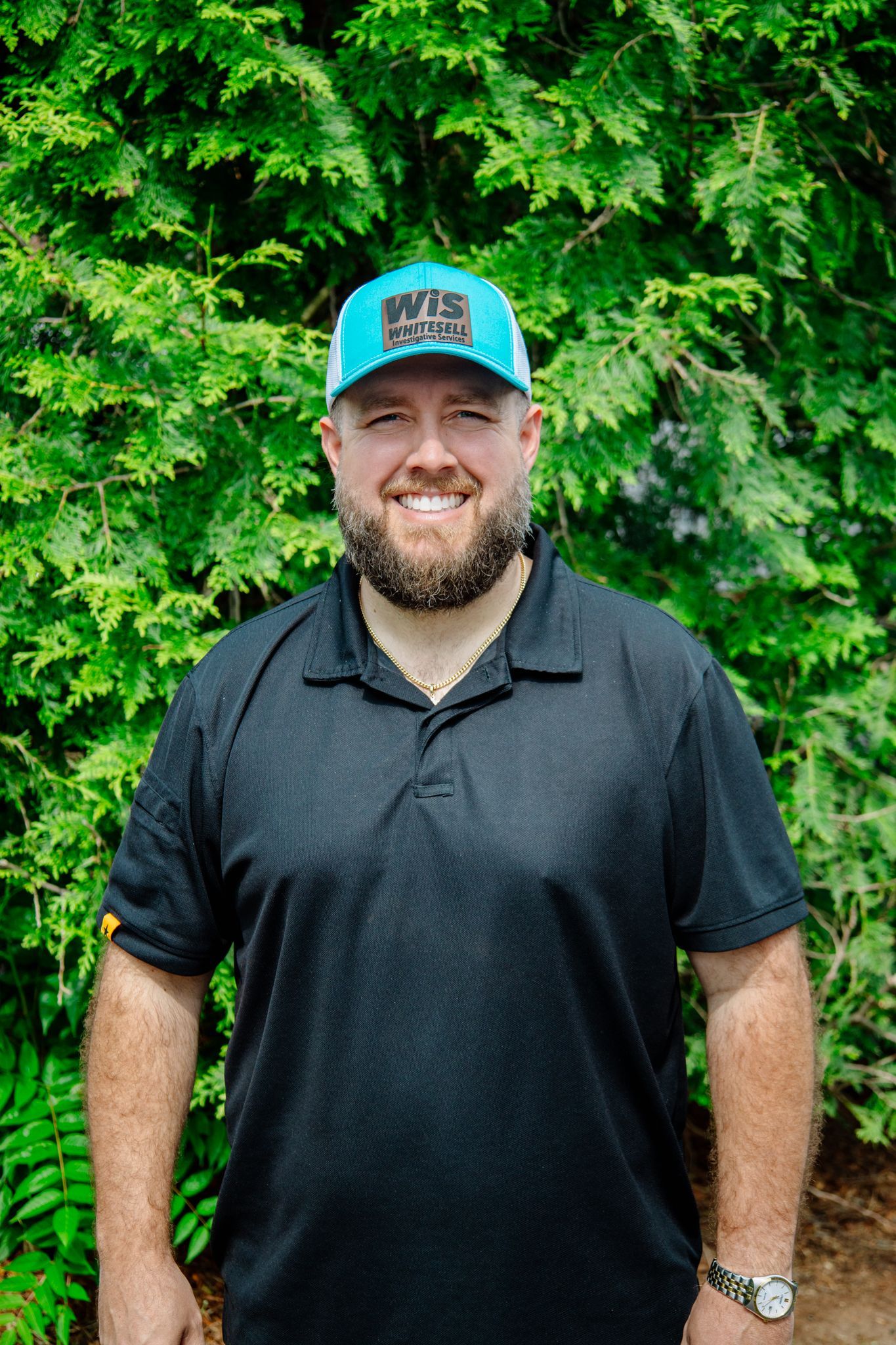 Man with beard wearing a blue cap and black polo, smiling in front of green foliage.