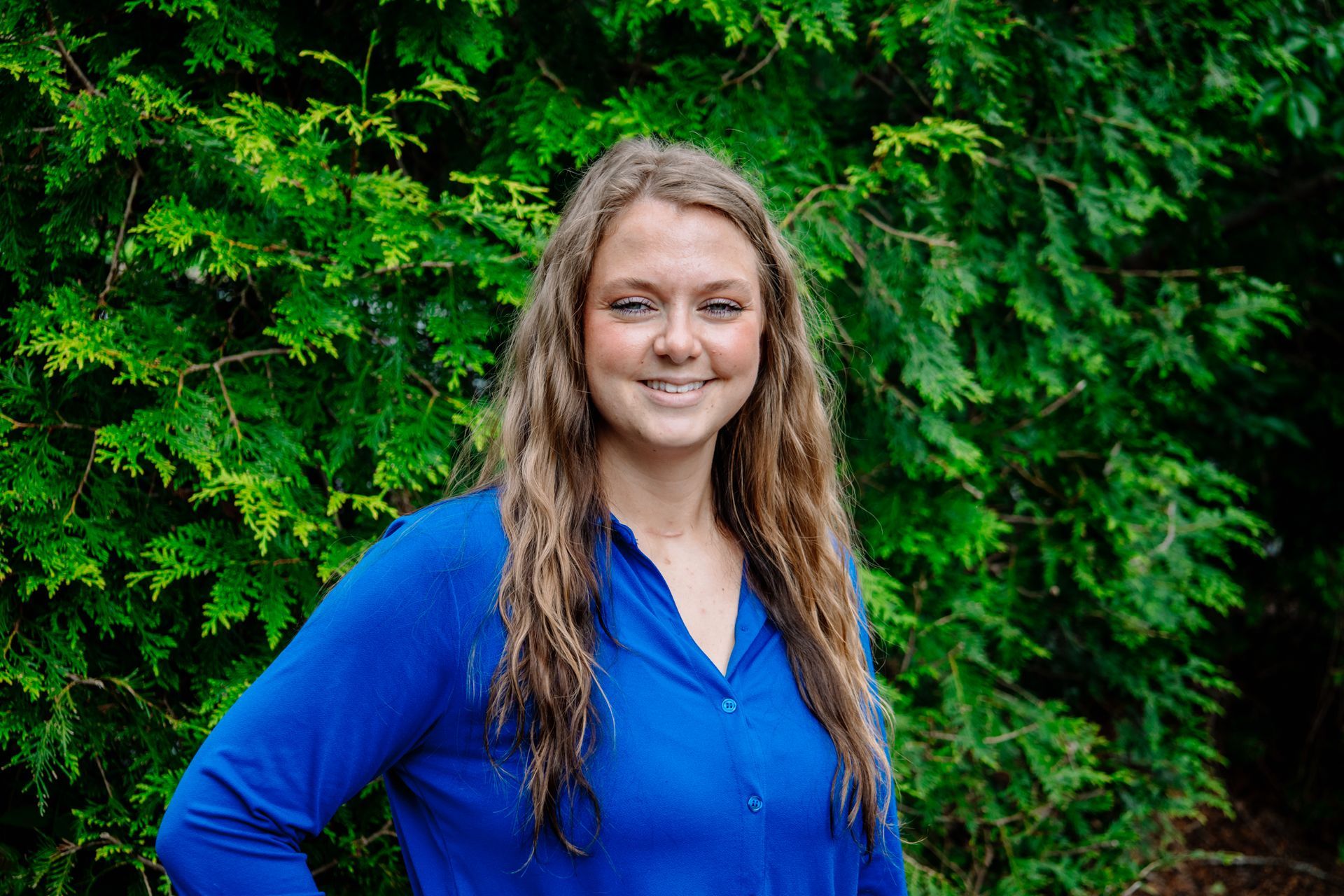 Woman with long brown hair smiles, wearing a blue shirt, standing in front of green foliage.
