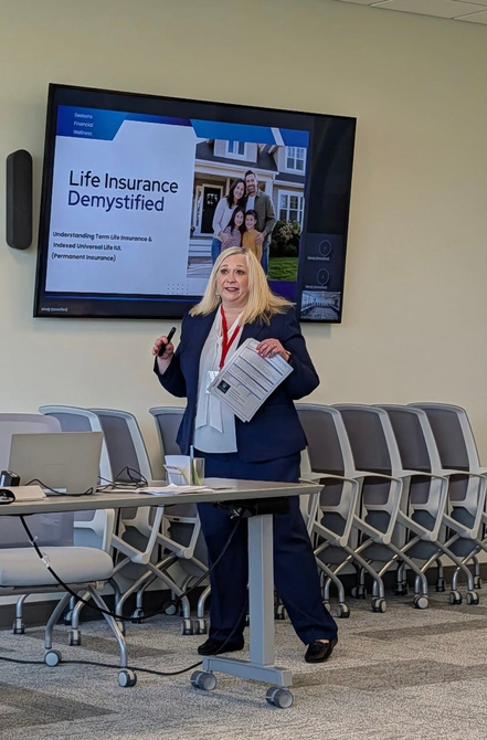 Speaker at podium in a conference room, standing beside rows of folding chairs and a presentation screen.