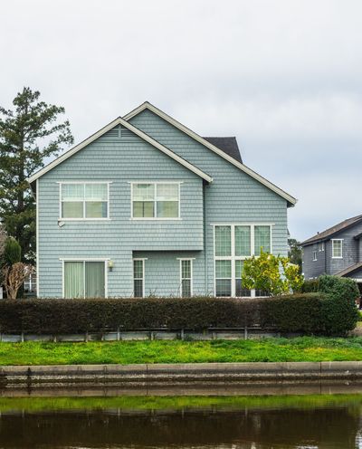 Blue-gray house with white trim behind a hedge, viewed across a canal under a cloudy sky