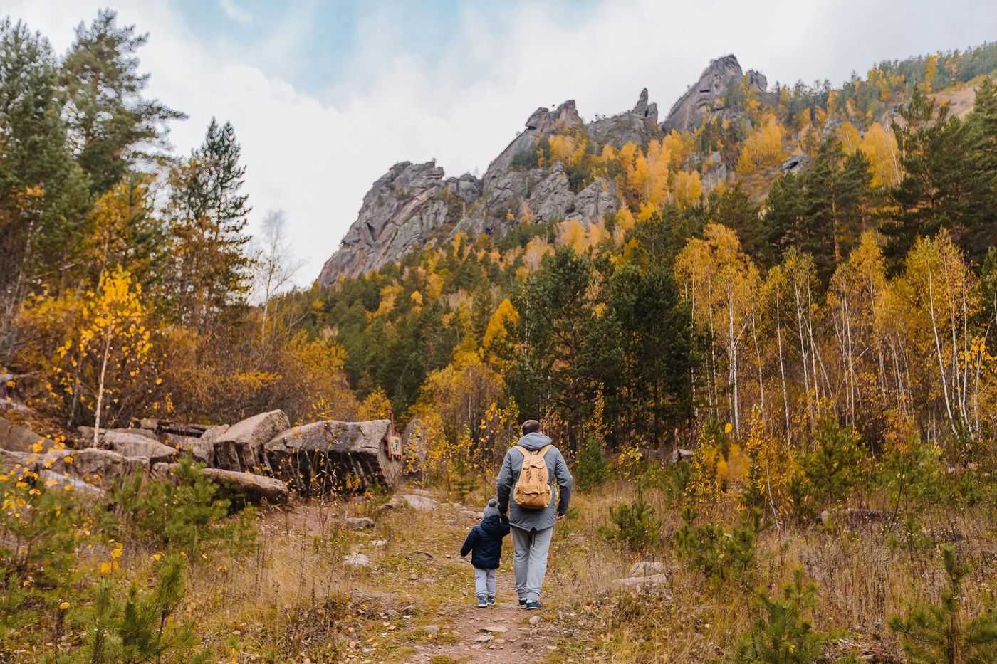 Two hikers on a trail beside yellow autumn trees and rocky mountains under a cloudy sky