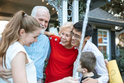 Smiling family huddled together outdoors in front of a house, with a child holding a yellow shopping bag