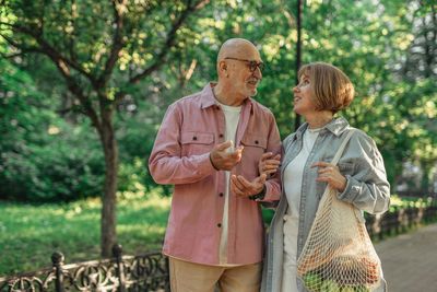 Two people chatting on a tree-lined path, one in a pink jacket and one holding a white bag.