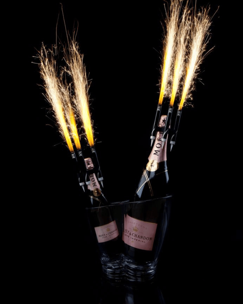 Two champagne bottles with sparklers, in glasses against a black background.