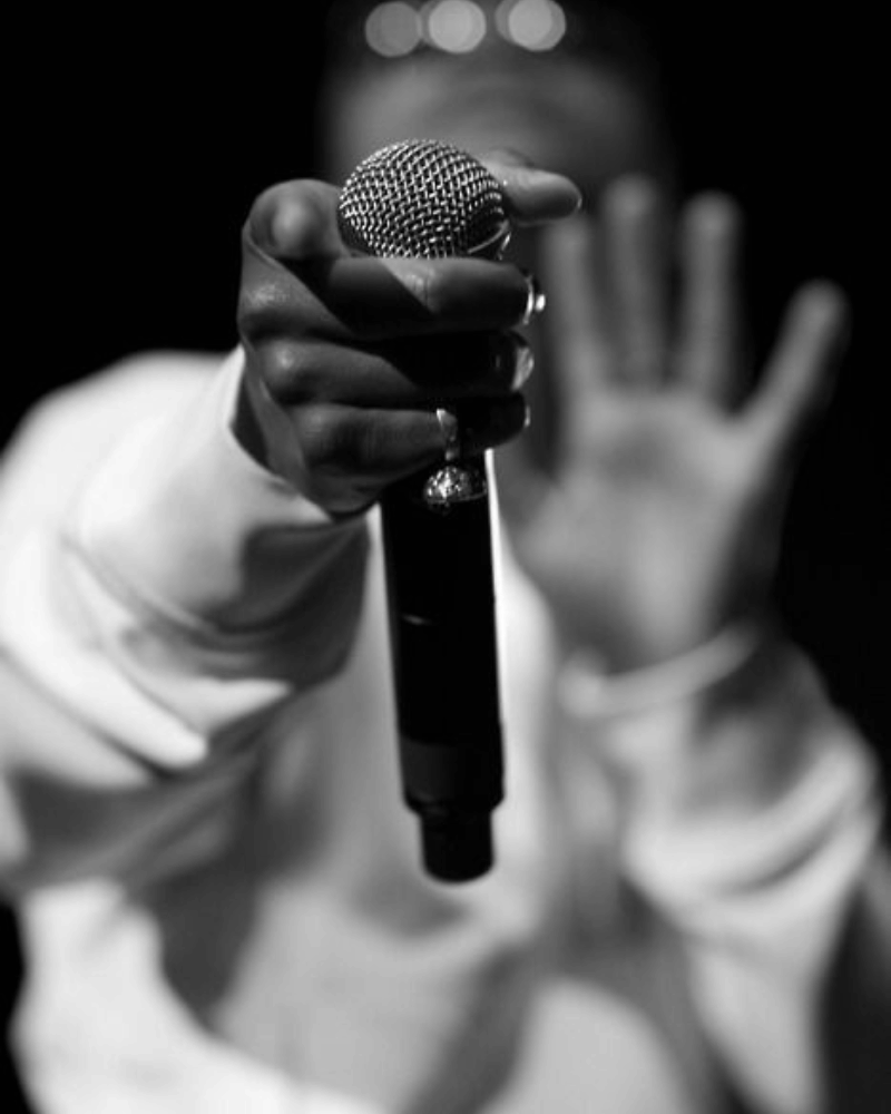 Person's hand gripping a microphone, other hand raised with fingers spread out, on a dark stage.