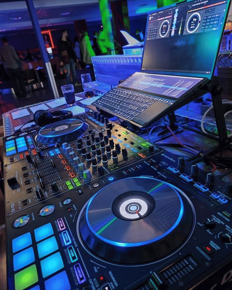 DJ setup with laptop displaying waveforms, controllers, and microphone in a dimly lit club.