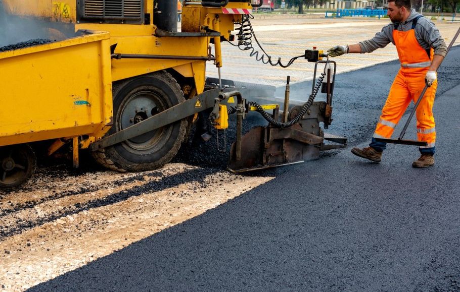 Road worker in orange gear operating asphalt paver, laying fresh black pavement beside a yellow machine.
