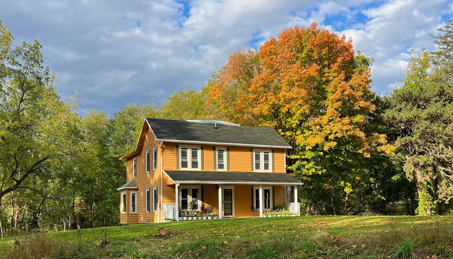 A yellow house with a porch in the middle of a field surrounded by trees.