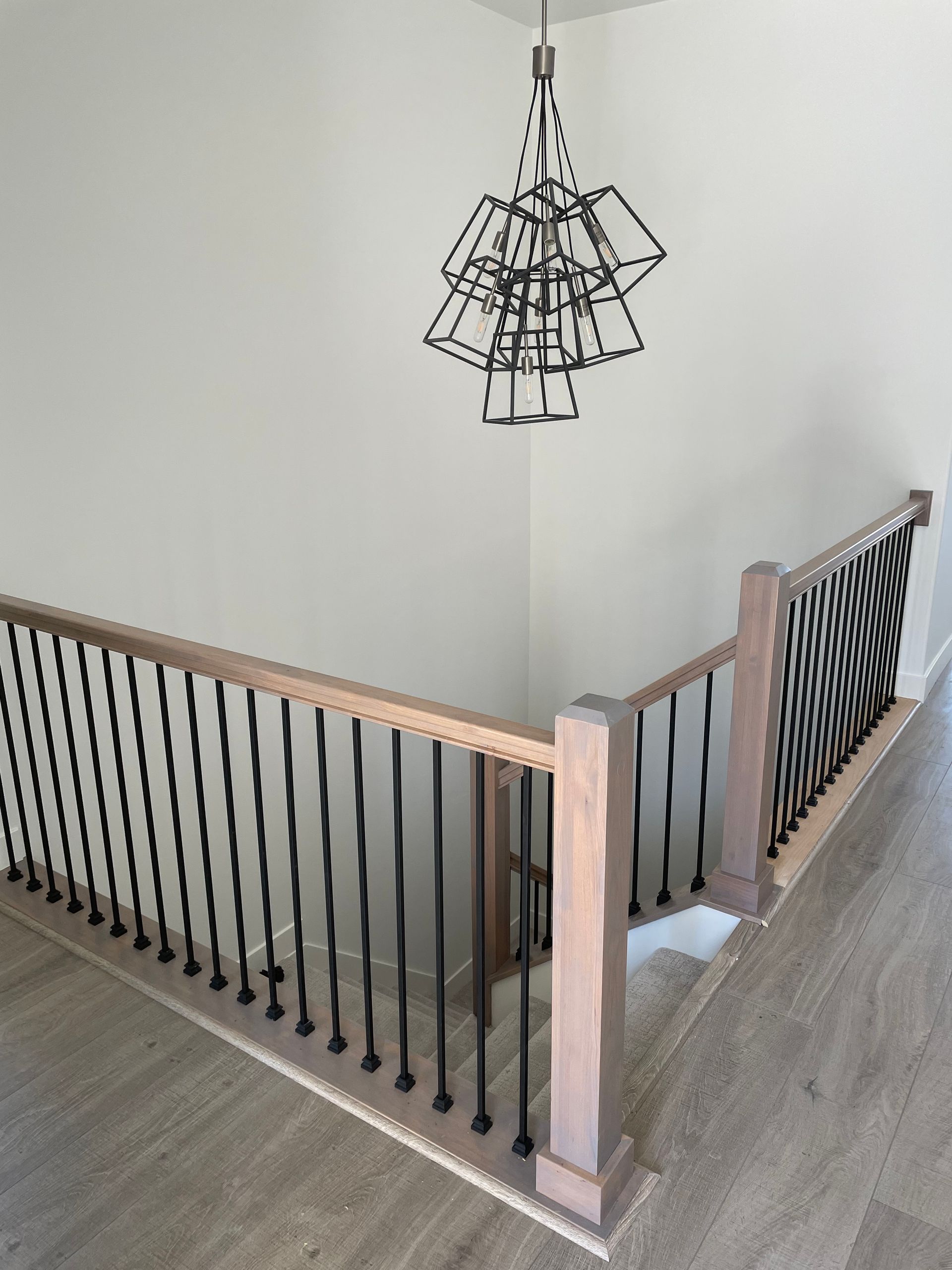 Staircase with wooden handrails, black iron balusters, and a modern chandelier; light-colored flooring and walls.