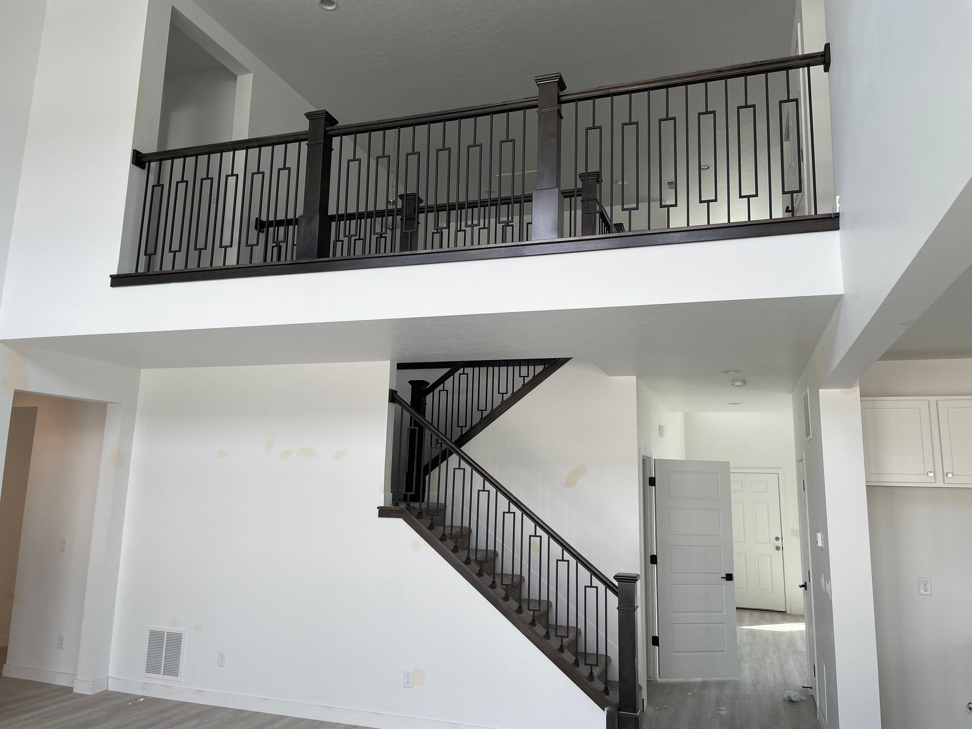 Two-story house interior with a staircase and a balcony with patterned black railings. White walls.