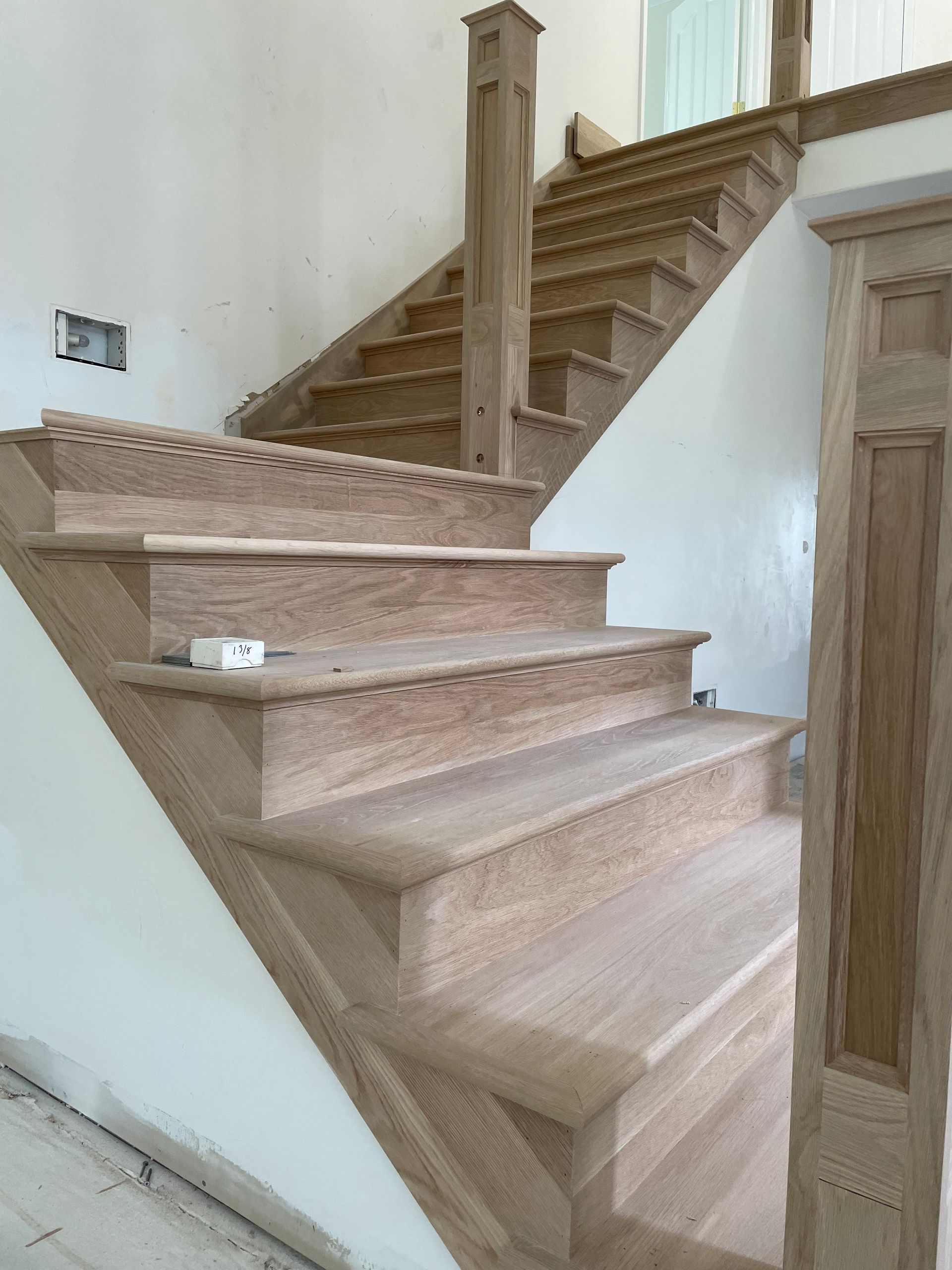 Unfinished wooden staircase with decorative edging and posts. Staircase is against a white wall.