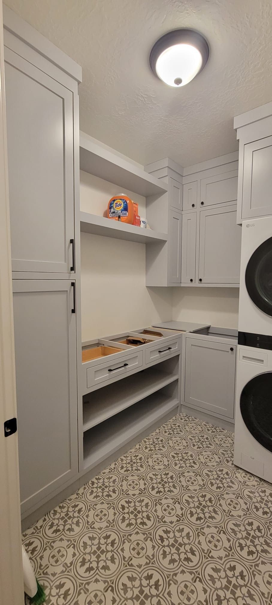 Laundry room with gray cabinets, shelves, and patterned floor. Stacked washer and dryer.