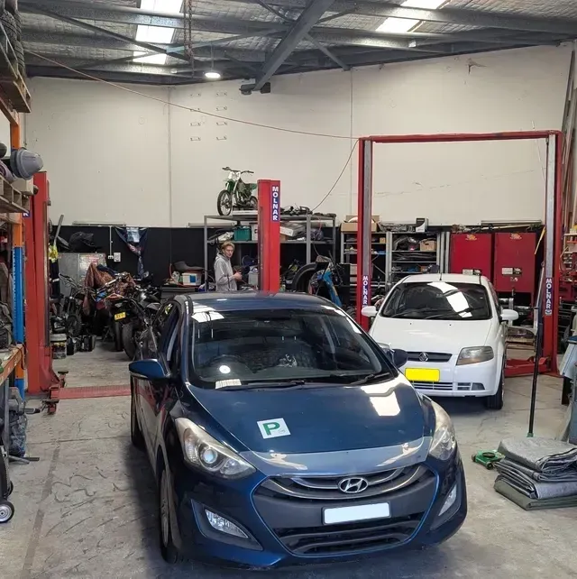 Inside a car repair shop: blue car in front, white car behind, mechanics and tools visible — ATD Automotive in North Wollongong, NSW