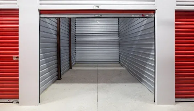 An empty, clean storage unit with light grey metal walls and floor, flanked by closed red roll-up doors.