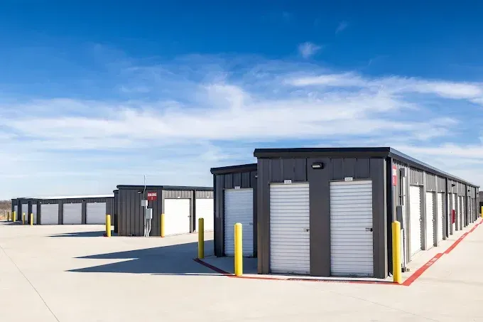 Rows of dark grey self-storage units with white roll-up doors on a concrete lot under a bright blue sky.