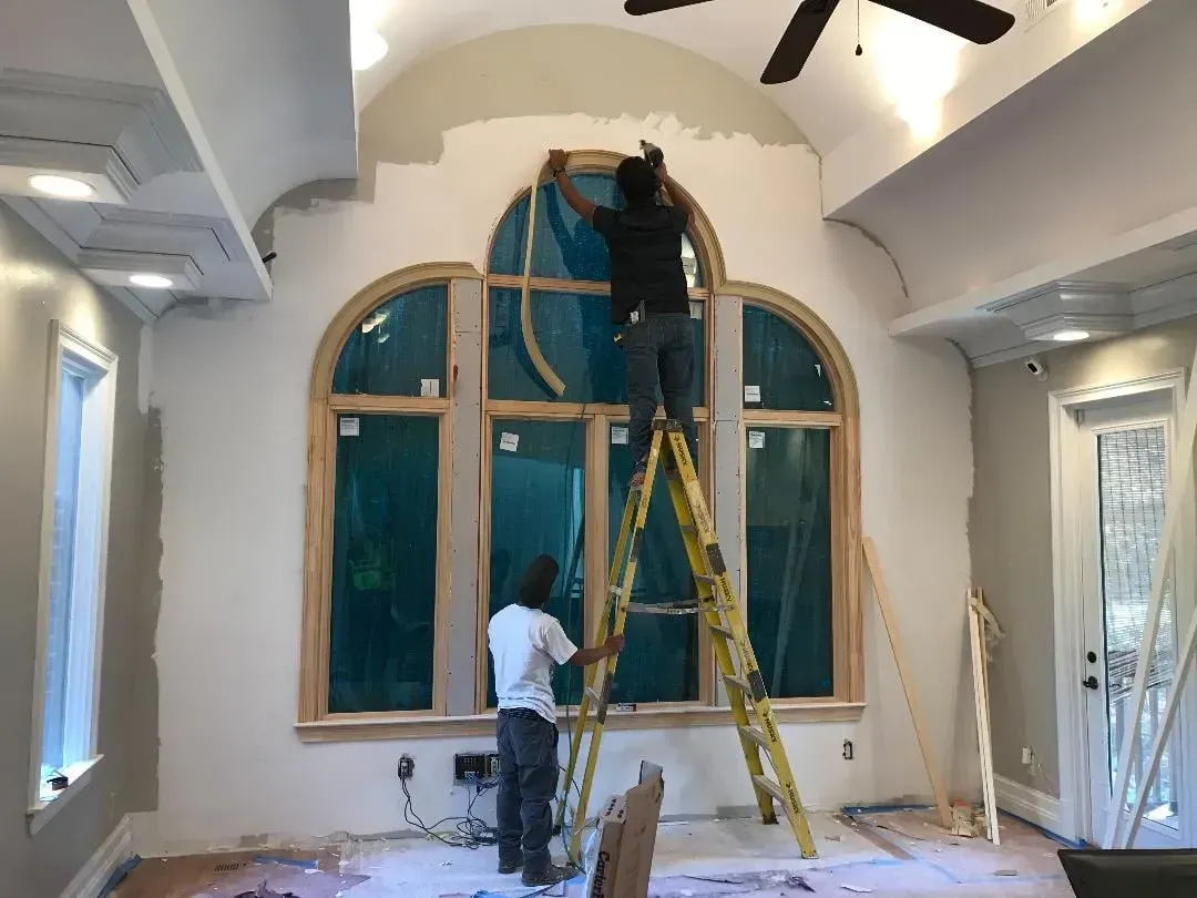 Two men are painting a large window in a living room.