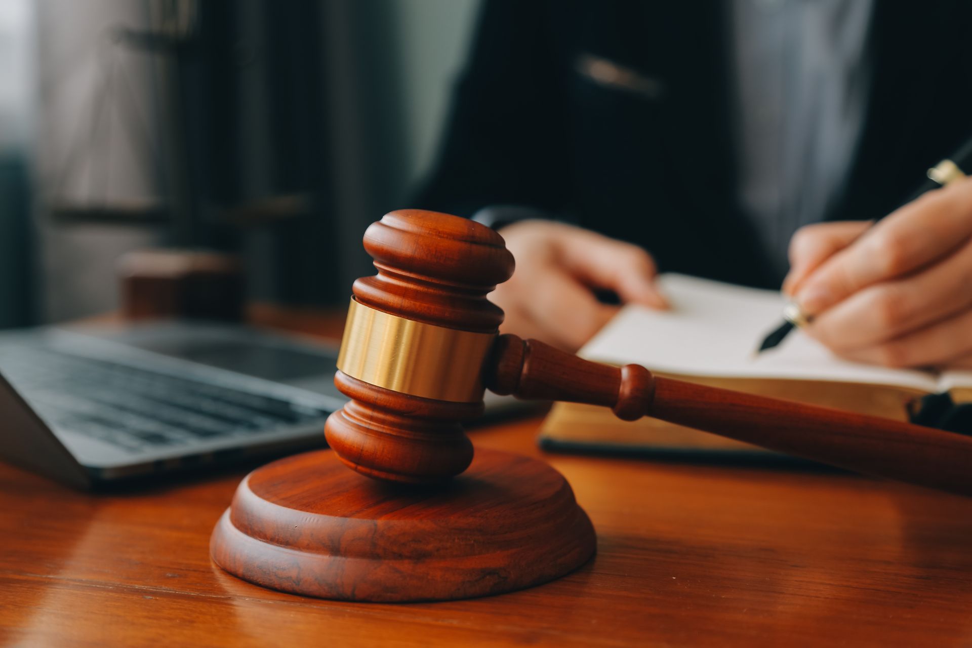 A judge 's gavel is sitting on a wooden table next to a person writing in a notebook.