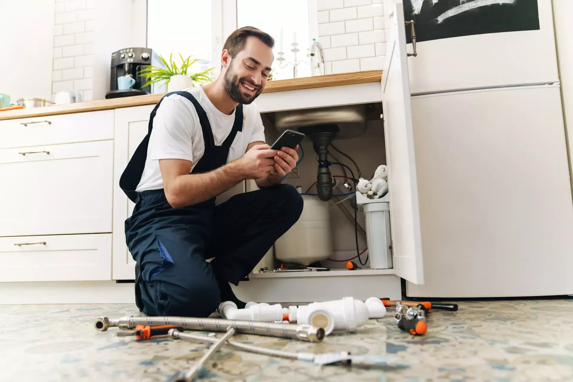 A plumber is kneeling on the floor in a kitchen looking under a sink.