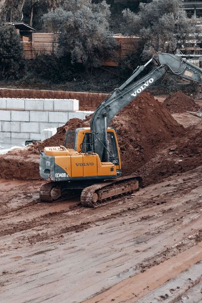 Yellow excavator on a muddy construction site beside a large dirt pile and retaining wall