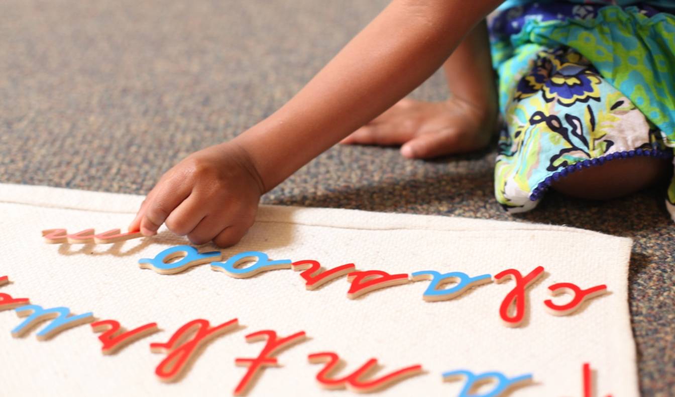 Child's hand arranging blue and red cursive letters on a white mat.
