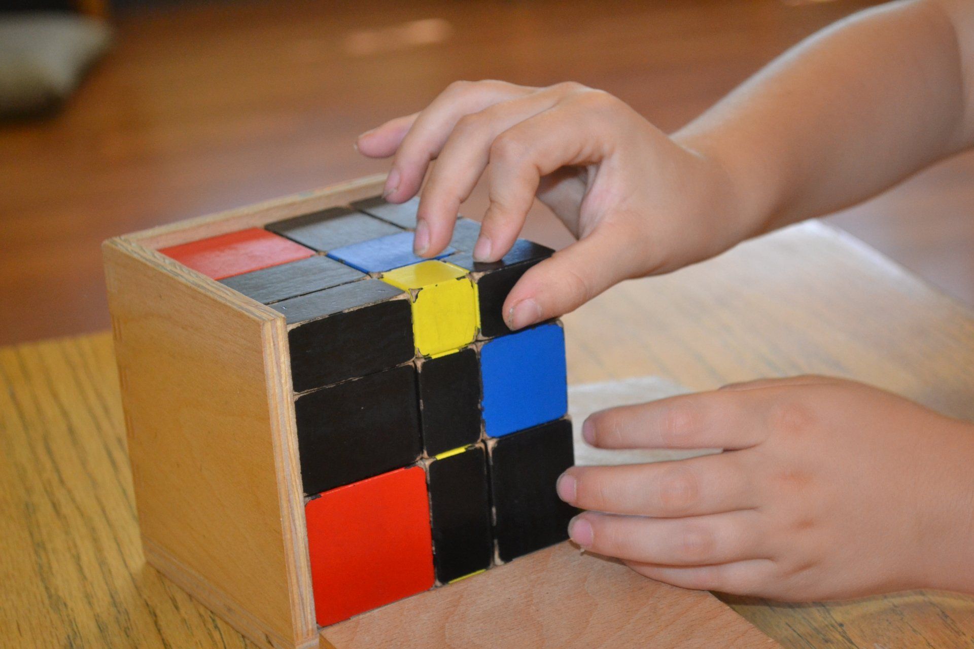 Child's hands assembling a wooden cube puzzle with colored blocks: red, blue, yellow, and black.