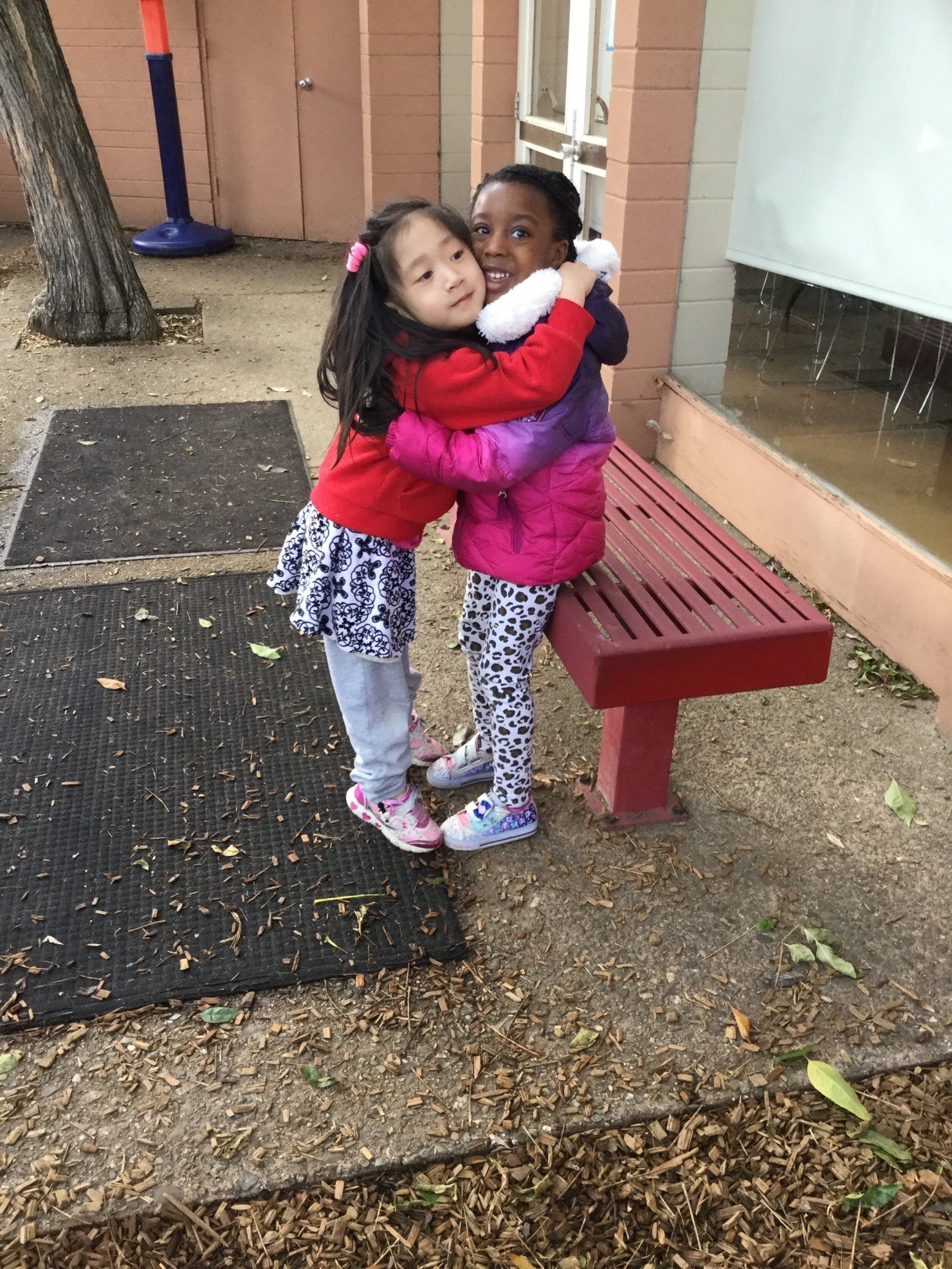 Two young girls, one Asian, one Black, hugging on a bench outdoors.
