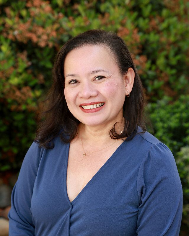 Smiling woman in a blue top, brown hair, outdoors against green foliage.
