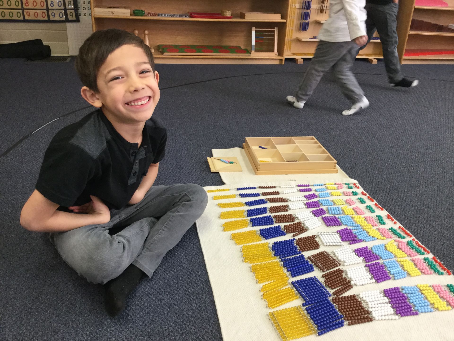 Young girl with long dark hair wearing pink jacket, seated at wooden desk, writing on a worksheet with pencil.