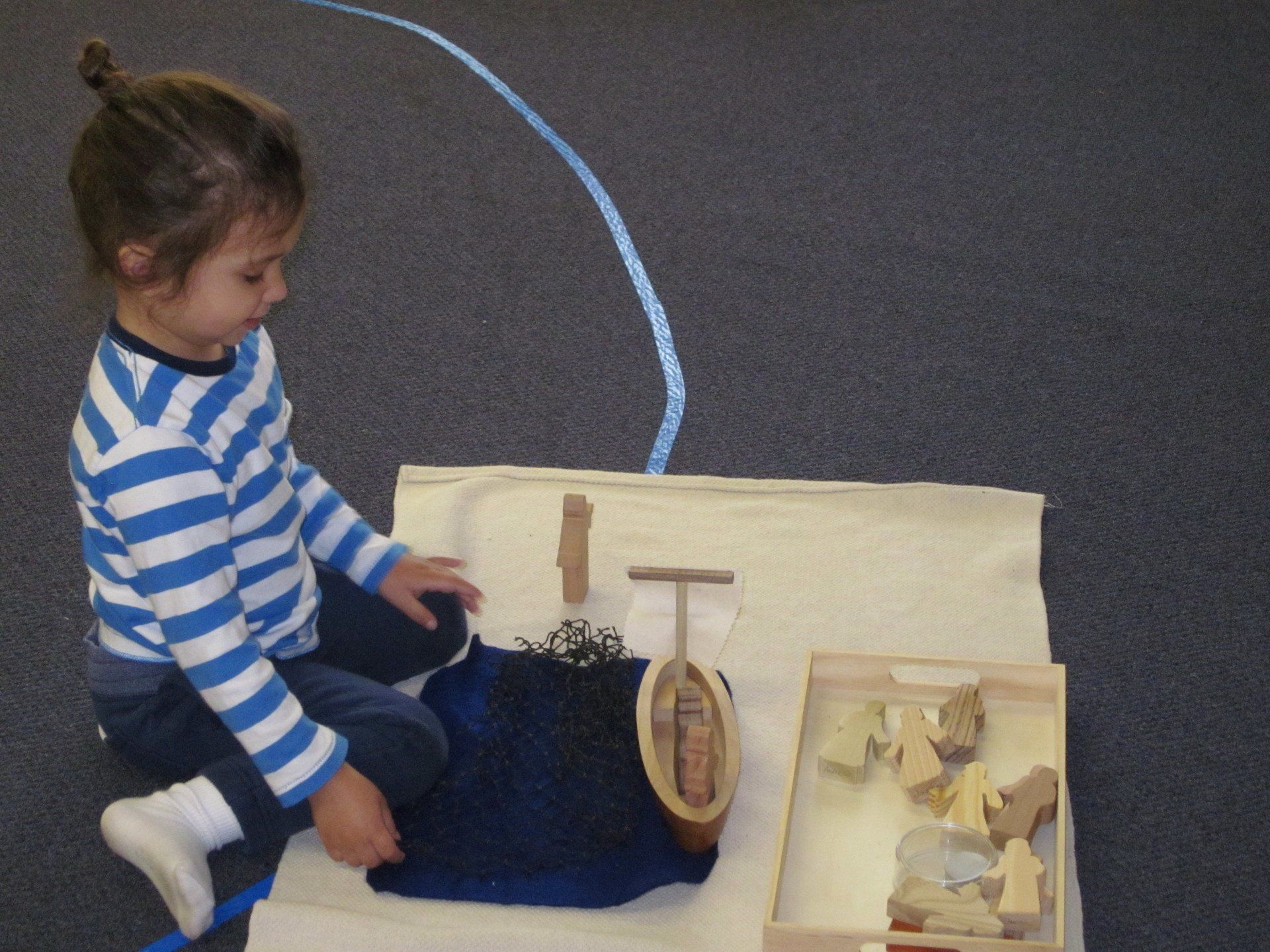 Young child seated, playing with a wooden boat and figures on a cloth, blue floor, focused.