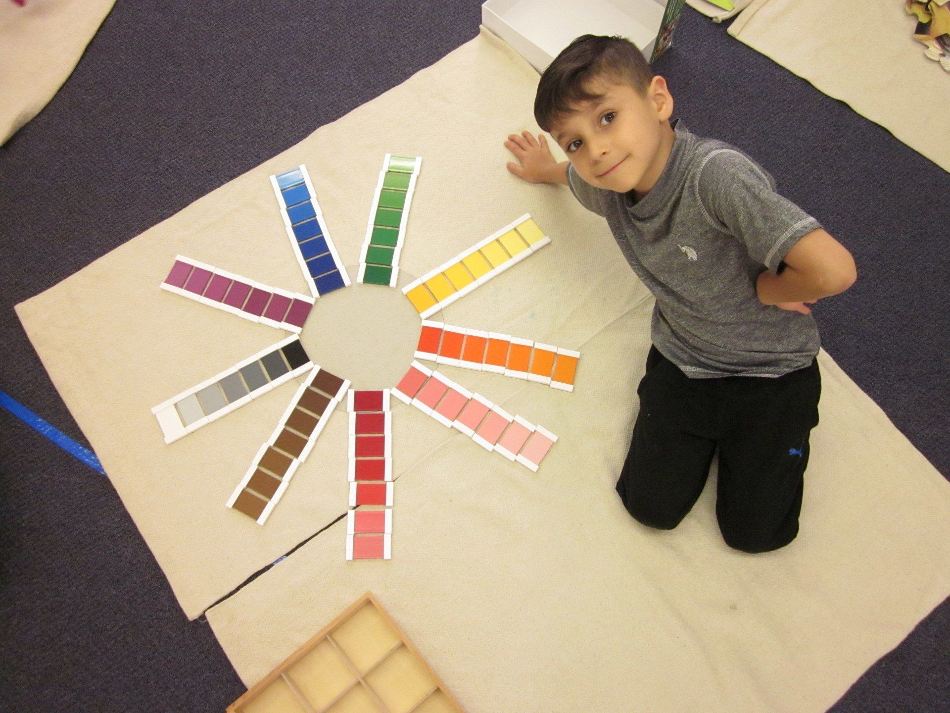 A young boy kneels, displaying a color wheel made of gradient-colored rectangles on a rug.