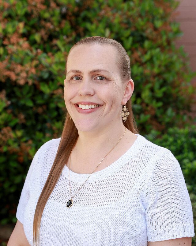 Woman with long blonde hair smiles, wearing a white sweater and necklace, outdoors in front of green foliage.