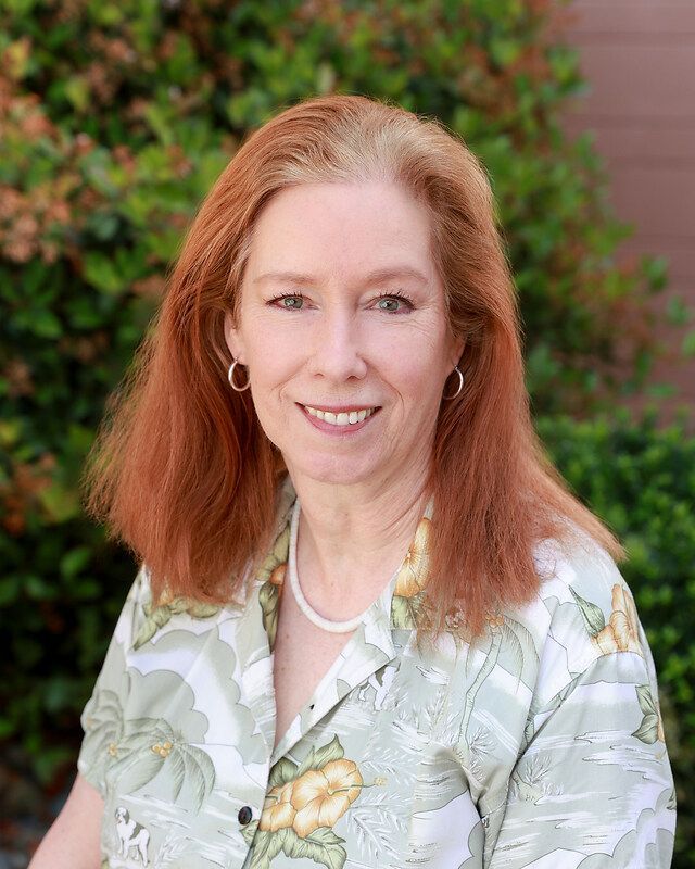 A woman with red hair is wearing a blue shirt and a purple vest.