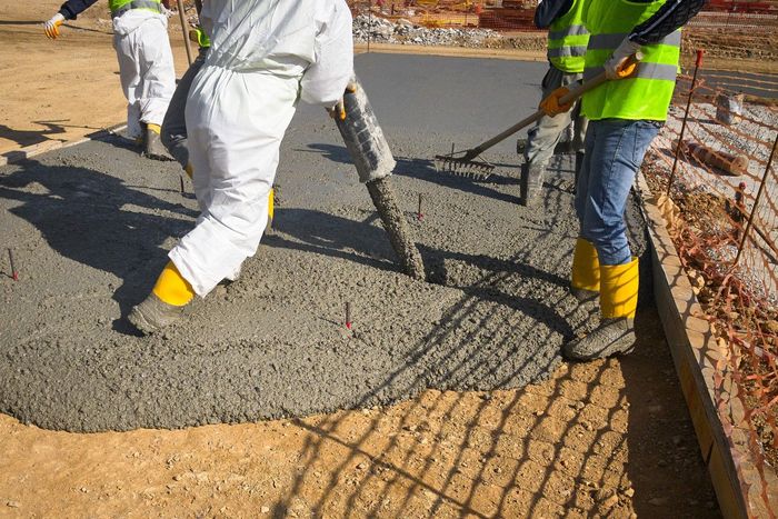 Construction workers pouring and leveling wet concrete on a construction site. One worker wears a protective suit and boots, while another wears a safety vest.