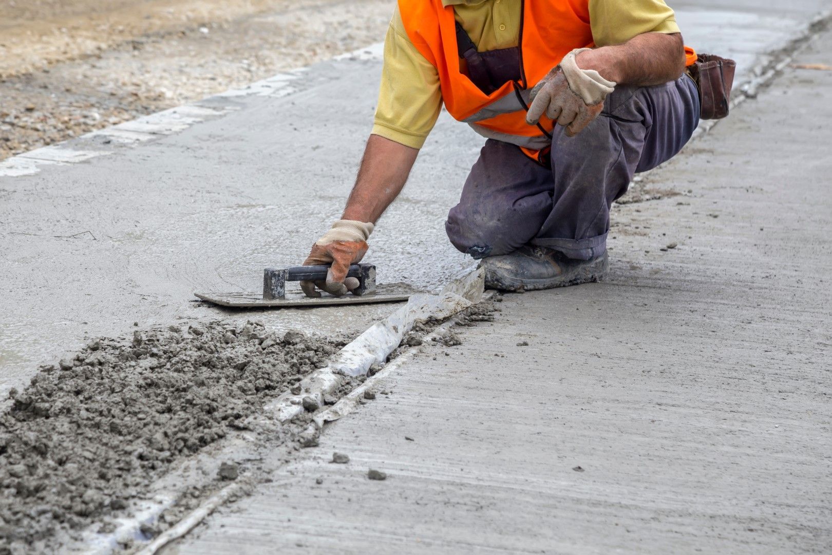 Construction worker kneeling, using a trowel to smooth concrete in a road joint, wearing orange vest and gloves.