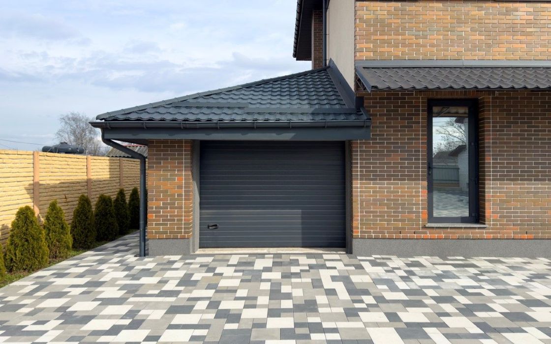 Garage with gray door, brick facade, and patterned pavers. Black roof with gutter.