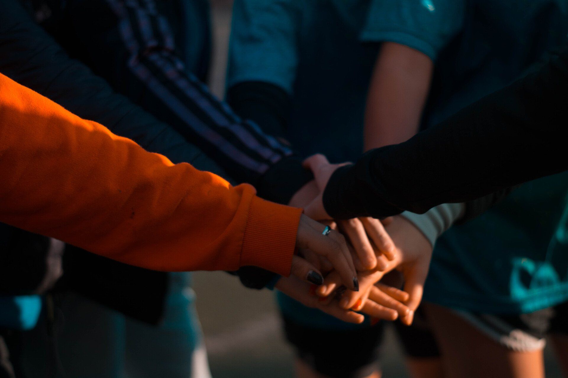 Hands stacked together in a sports team huddle, with orange and teal jerseys visible.