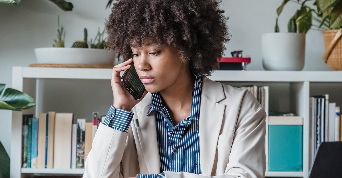 Person on the phone at a desk in front of a bookshelf, wearing a beige blazer and blue striped shirt.