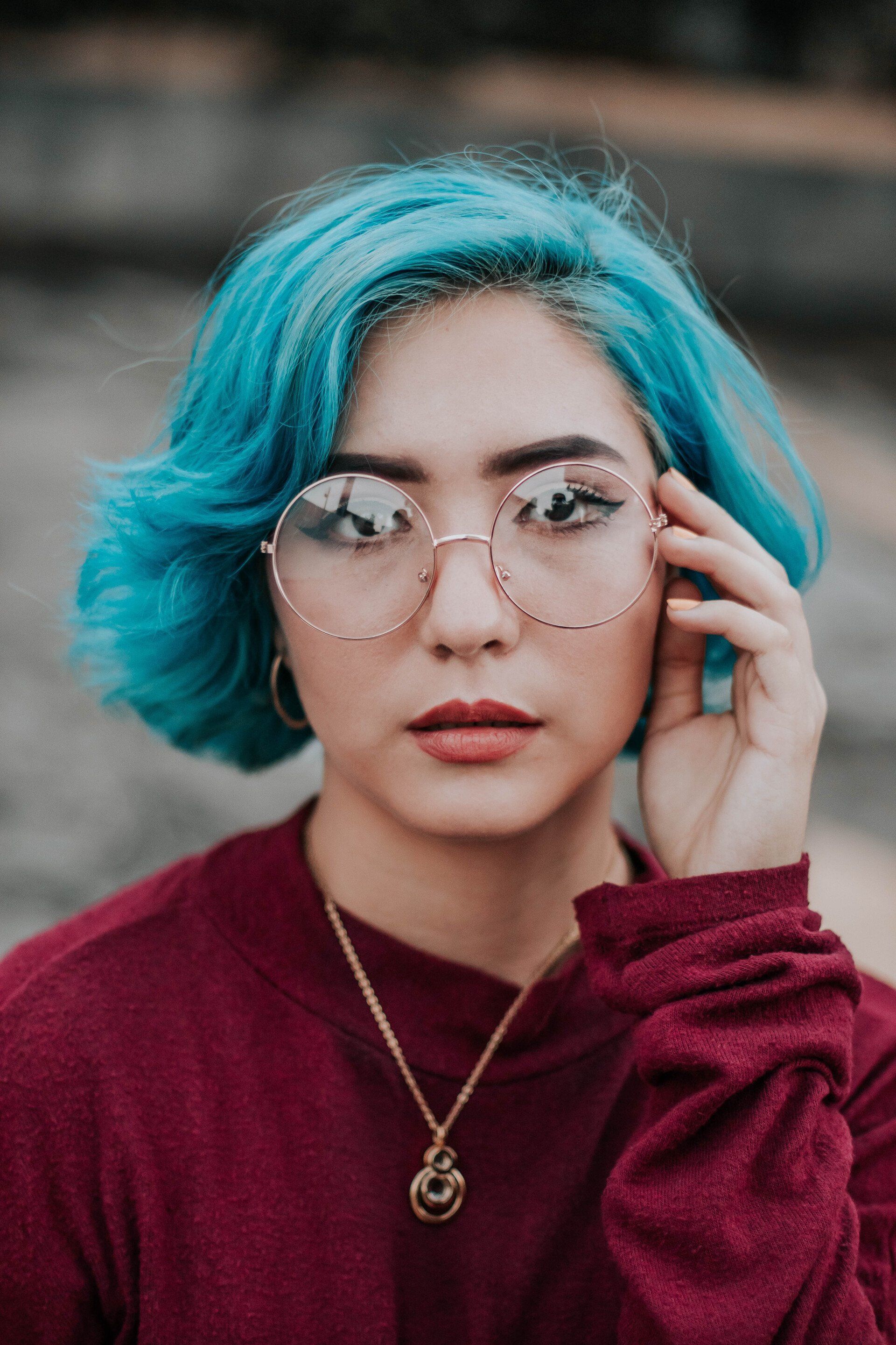 Person with bright blue hair and glasses, wearing a burgundy top, touching their face outdoors.