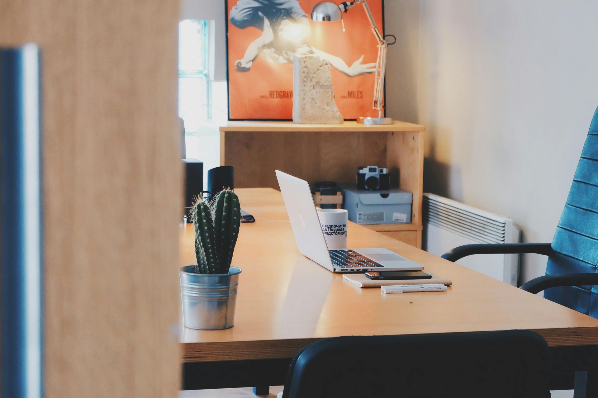 A clean, modern home office desk with a laptop, a small potted cactus, a desk lamp, and an orange wall poster.