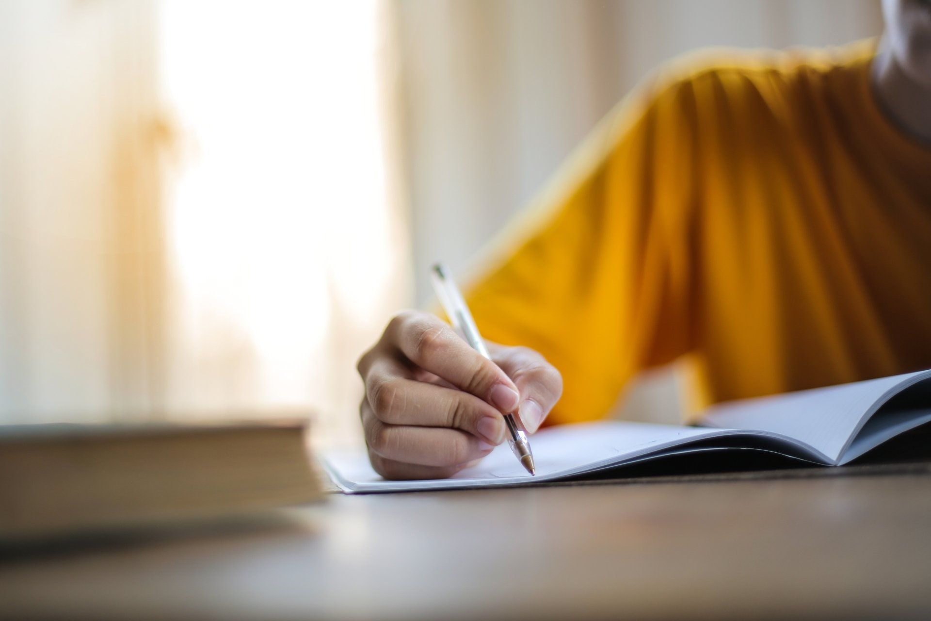 Person in a yellow shirt writing in an open notebook on a table indoors