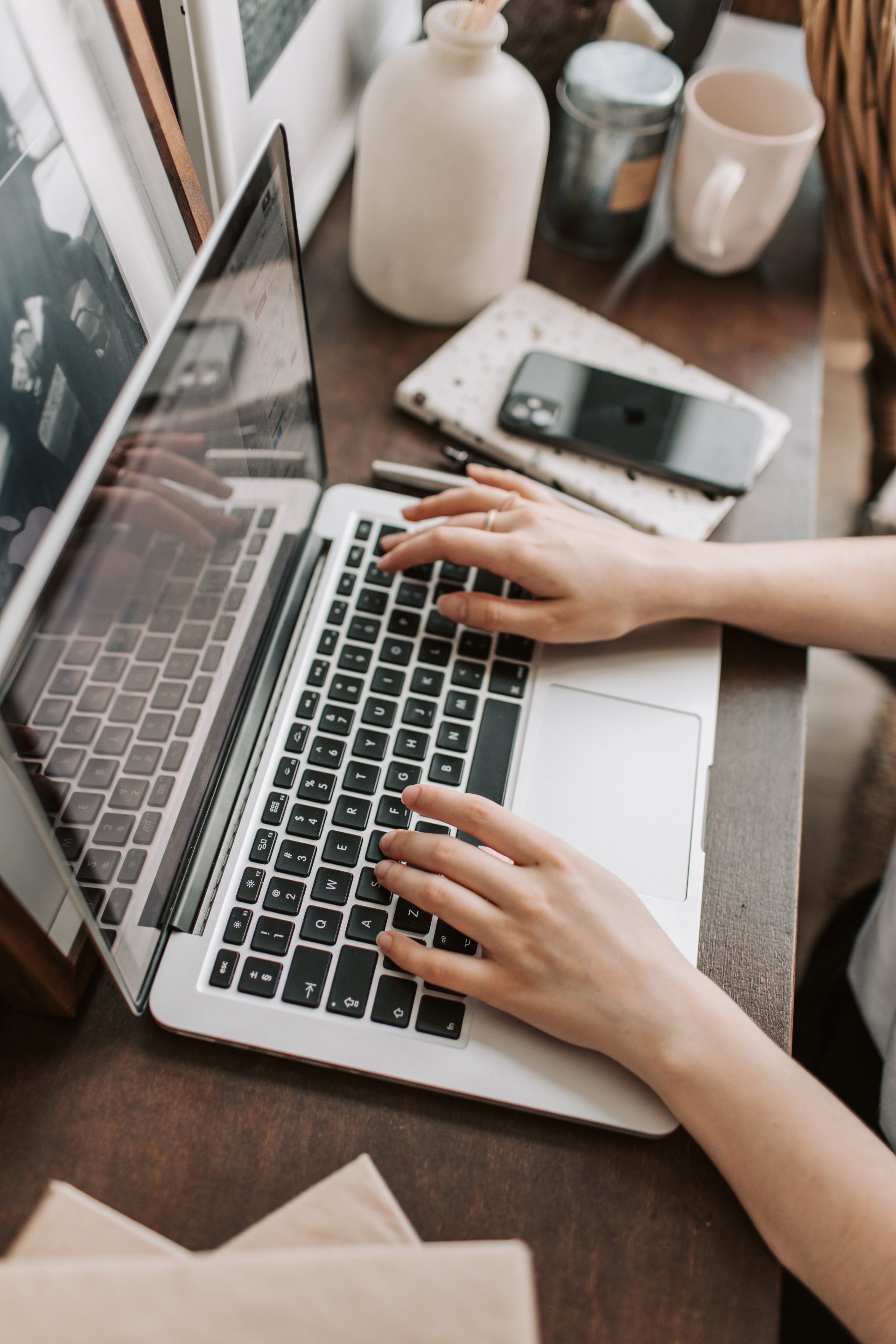 Hands typing on a laptop beside a smartphone on a wooden desk with a mug and vase.