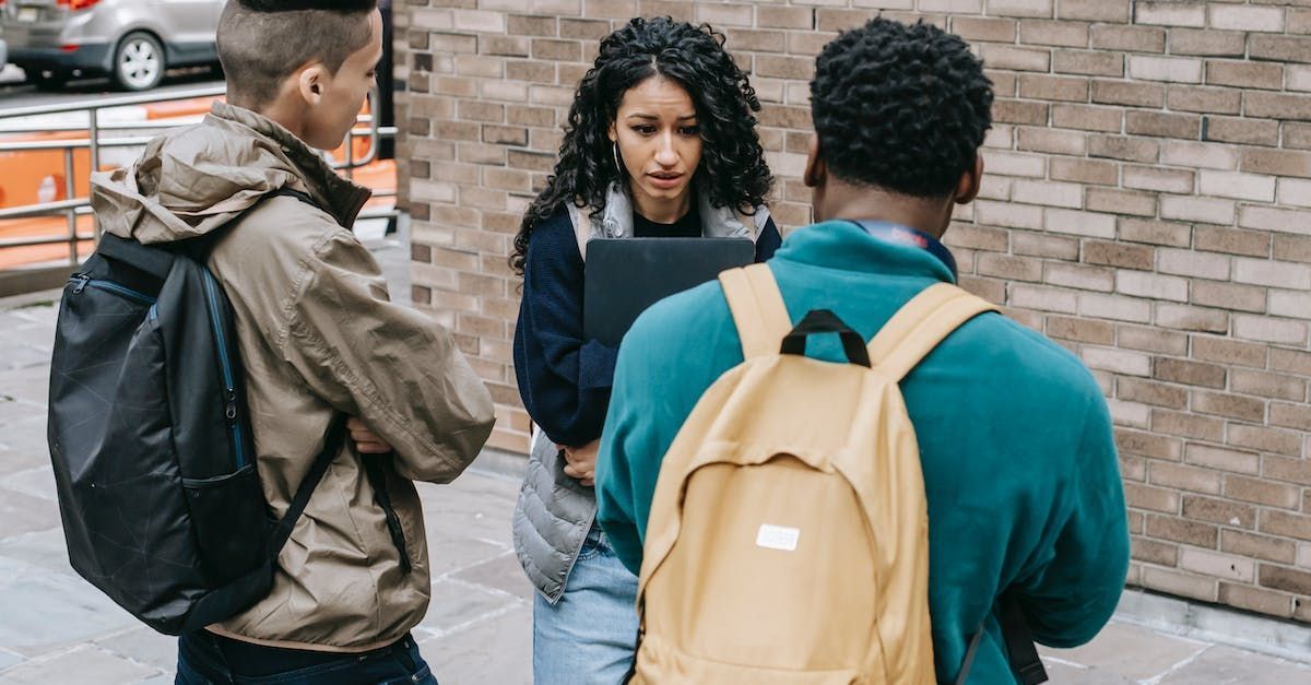 Three students talking on a sidewalk, one holding papers, with backpacks near a brick wall.
