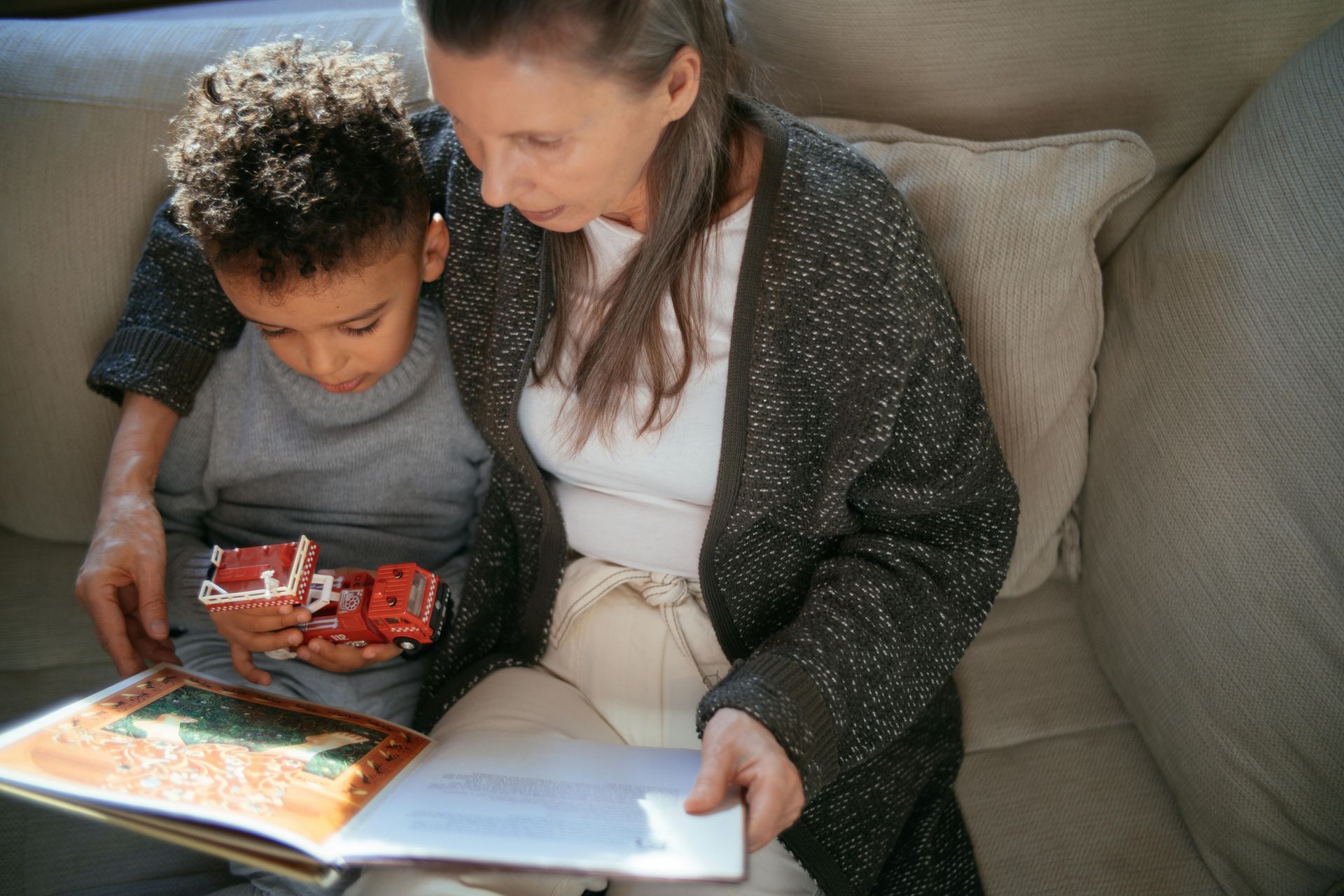 An adult sits on a sofa, reading an open book to a child holding a red toy car.