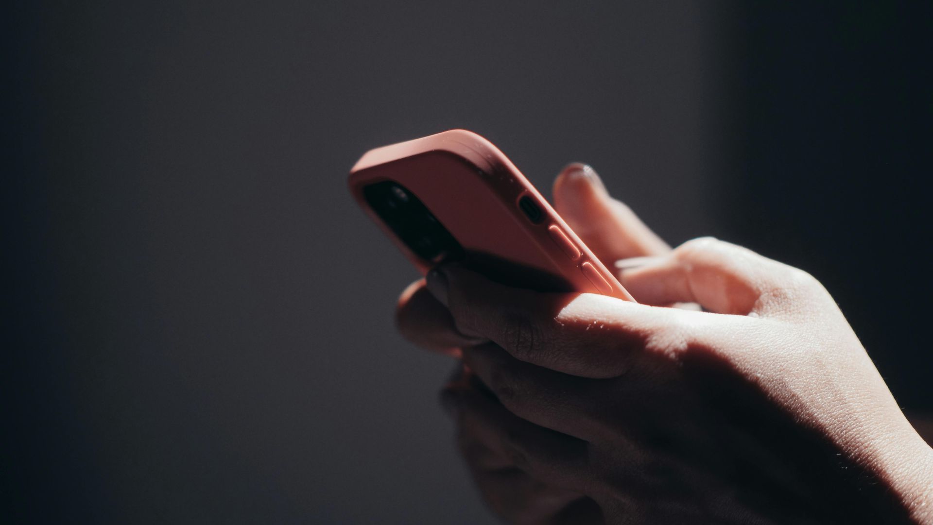 A person holds a salmon-colored smartphone, their hands illuminated against a dark, moody background.