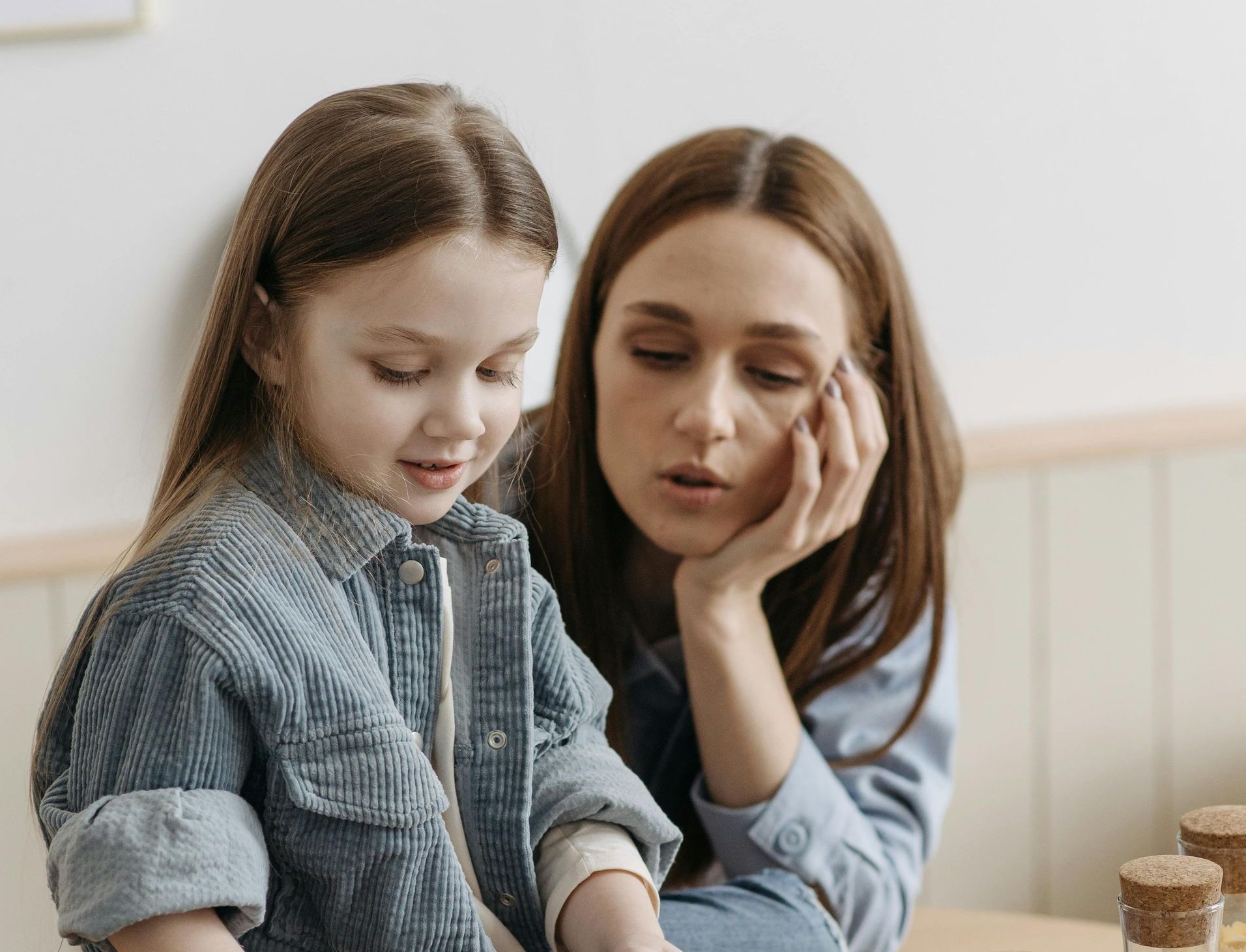 Woman leaning toward smiling child, both focused on something on a table in a bright room.
