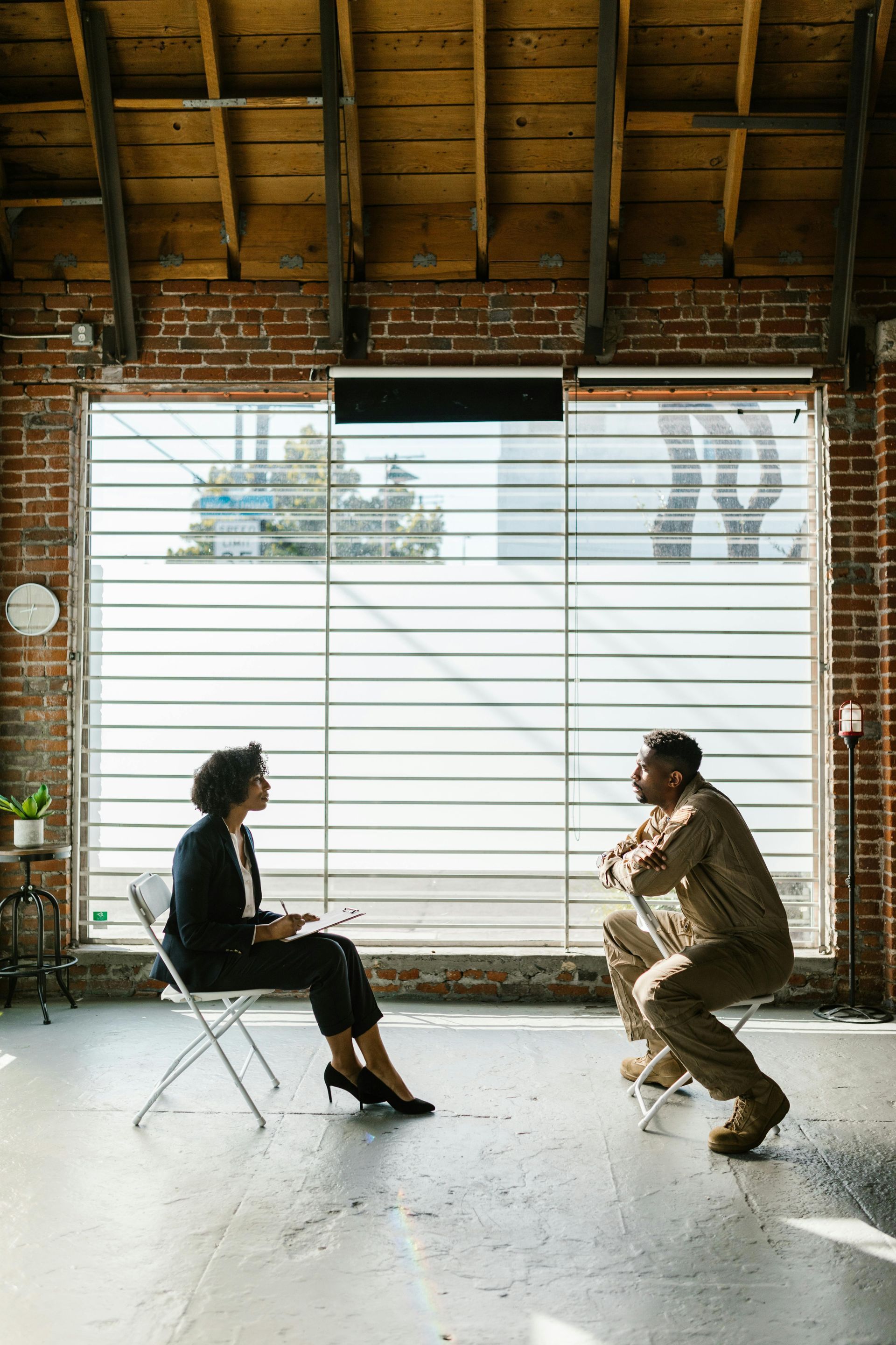Two people seated facing each other in a bright loft with brick walls and a large window