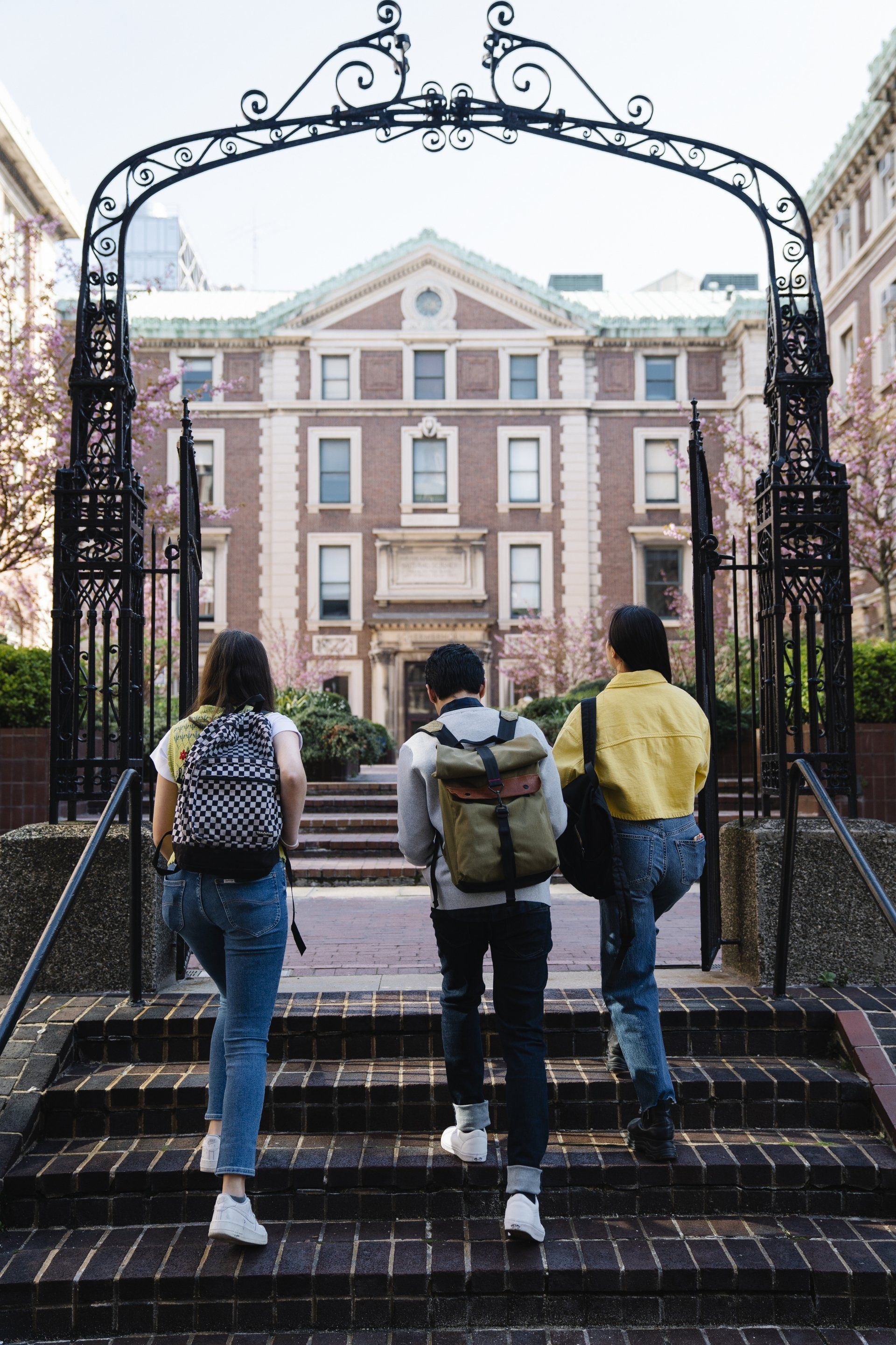 Three people walking up steps through an ornate black gate toward a historic brick building