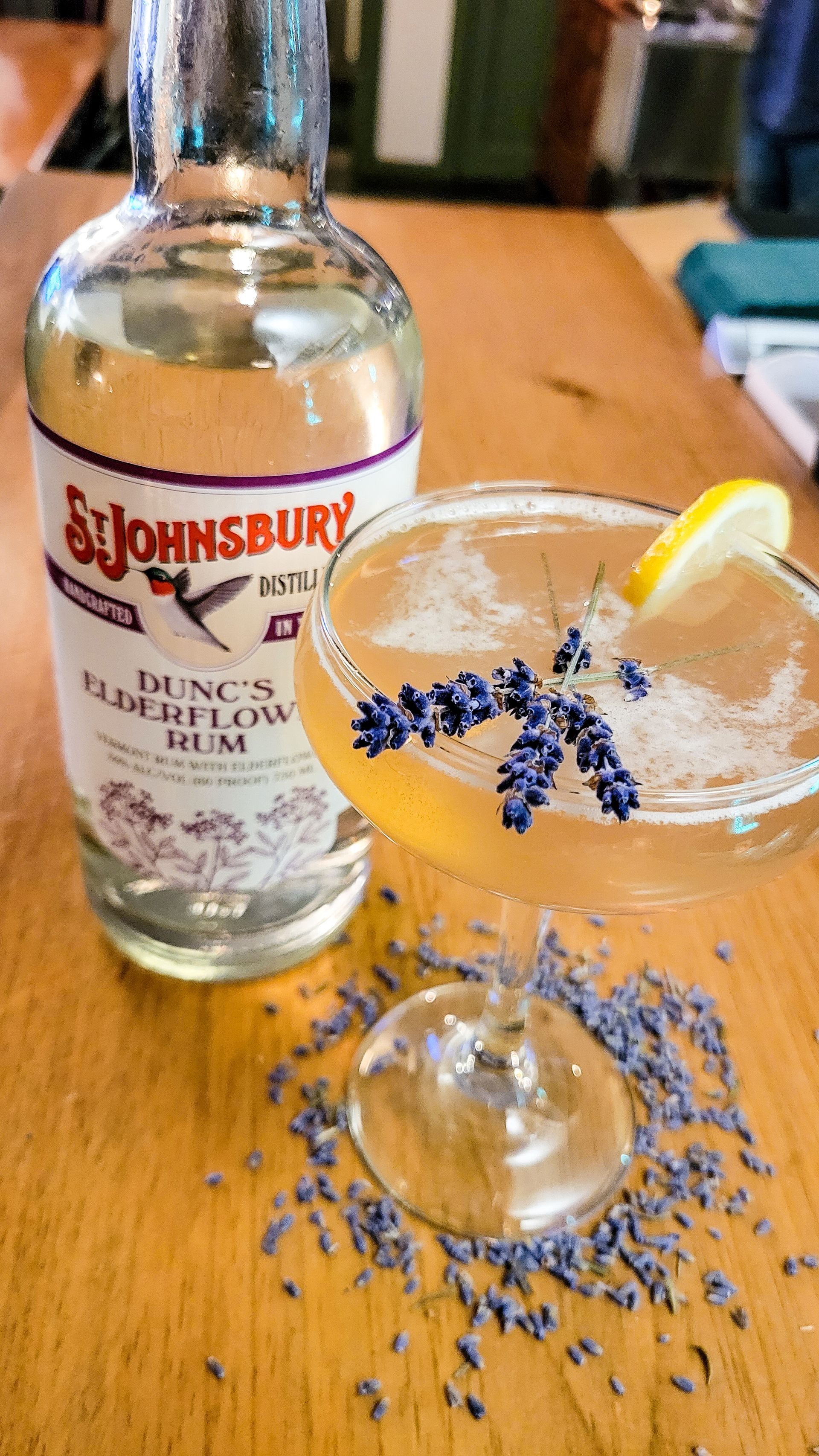 A bottle of St. Johnsbury distillery Dunc's Elderflow Rum. With a glass full of Rum, a slice of lemon and a decorative spring of Lavender.